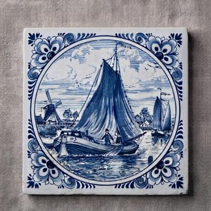May include: A square ceramic tile featuring a blue and white hand-painted scene of a sailboat on water with a windmill in the background. The central circular illustration is framed by ornate floral corner motifs in a classic delft blue style.