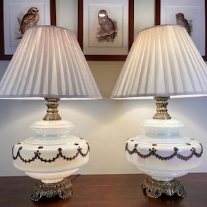 May include: Pair of vintage table lamps with white pleated shades, ornate brass bases, and iridescent white glass bodies. The lamps have a decorative black pattern around the middle. Framed owl prints are in the background.