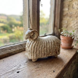 May include: A ceramic ram figurine with a cream-colored body and brown horns. The sculpture is on a wooden windowsill, with a blurred outdoor scene visible through the window. A small terracotta pot with a plant is also on the sill.