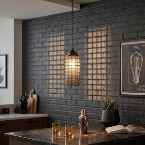May include: A cylindrical pendant light fixture featuring a textured, amber-tinted glass shade encased in a dark metal grid. The light hangs from a black chain and ceiling mount, casting a grid-patterned shadow against a dark gray brick wall.