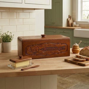 May include: A rectangular wooden bread box with a dark brown finish and a carved design featuring the text 'GIVE US THIS DAY OUR DAILY BREAD'. The box has a lid with a small knob handle. A glass butter dish with a wooden lid and a wooden knife are nearby.