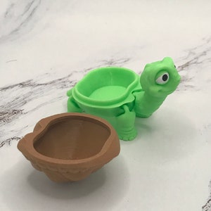 May include: A green turtle-shaped container with a brown shell-shaped lid. The turtle has a large eye and a small mouth. The container is made of plastic and is about 7.5 cm tall.