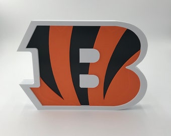 Pode incluir: Uma letra "B" impressa em 3D de cor branca, laranja e preta com um padrão de listras de tigre. A letra é um item decorativo.