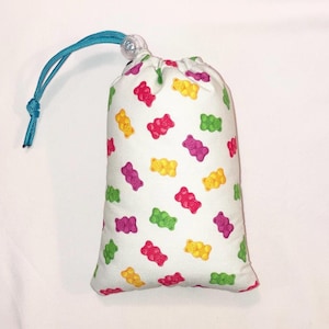 May include: A white drawstring bag with a teal drawstring and a gummy bear pattern. The gummy bears are in various colors, including red, yellow, green, and purple.