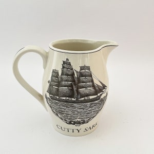 May include: White ceramic pitcher with a black and white illustration of a ship called "Cutty Sark" sailing on the ocean. The pitcher has a handle and a spout.