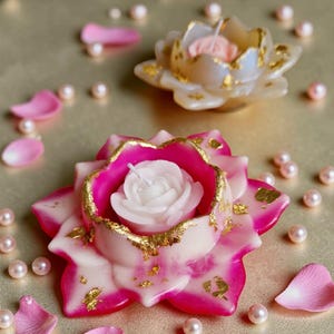 Op de afbeelding: Twee bloemvormige kaarsen met een witte roosvormige kaars erin. De kaarsen zijn roze en wit met goudbladaccenten. Verspreid liggen roze bloemblaadjes en imitatieparels. De achtergrond is een gouden oppervlak.