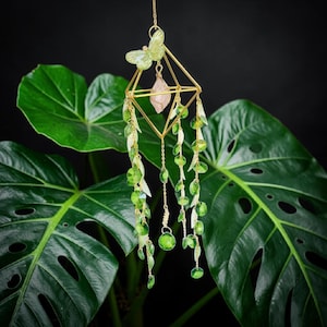 Puede incluir: Un atrapasoles colgante con un marco geométrico de metal dorado, un adorno de mariposa verde claro y un cristal rosa pálido central. Varias cadenas doradas cuelgan del marco, decoradas con pequeñas cuentas de cristal verde y dijes de hojas blancas.
