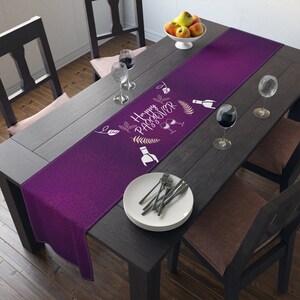 May include: Purple table runner with a white design that says "Happy Passover" with a bottle of wine, two wine glasses, and leaves. The runner is on a dark wood table with a plate and silverware.