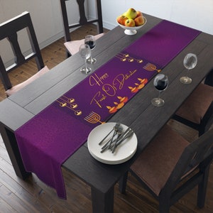 May include: Purple table runner with a gold design featuring the text "Happy Feast of Lights" and a menorah. The runner is on a wooden table with two wine glasses and a plate of silverware.