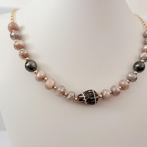 Puede incluir: Un collar de cuentas con cuentas facetadas de color marrón claro, cuentas esféricas lisas de color gris oscuro y una cuenta central de color marrón oscuro con un patrón. Las cuentas están separadas por pequeños espaciadores dorados.