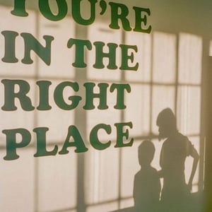 Op de afbeelding: Een witte muur met de tekst "You're in the right place" in groene letters. Twee silhouetten van mensen zijn zichtbaar in het raam achter de tekst.