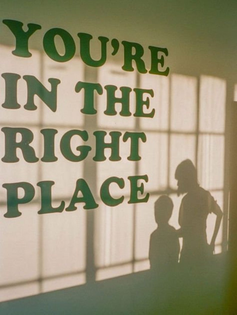 Op de afbeelding: Een witte muur met de tekst "You're in the right place" in groene letters. Twee silhouetten van mensen zijn zichtbaar in het raam achter de tekst.