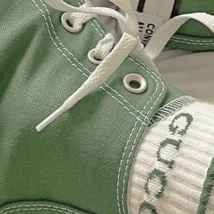 Op de afbeelding: Close-up van een paar groene canvas sneakers met witte veters en een witte sok met het woord "Gucci" erop geborduurd.