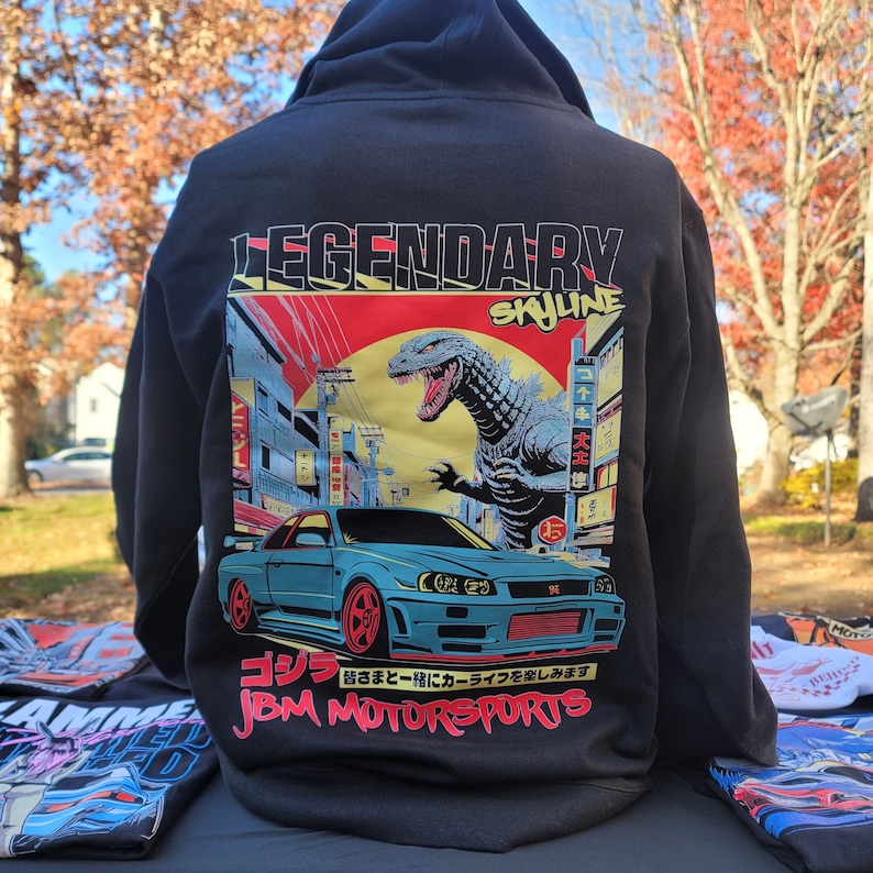 May include: Black hoodie with a graphic print of a blue car, a Godzilla-like monster, and Japanese text. The text on the hoodie reads "LEGENDARY SKYLINE" and "JBM MOTORSPORTS".