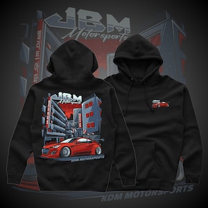 May include: Black hooded sweatshirt featuring a red sports car graphic with the text "JBM Motorsports" and "KDM Motorsports." The design includes a cityscape with red and gray buildings. A smaller car graphic is on the front.