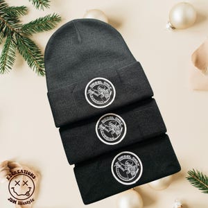 May include: Three dark gray and black beanies stacked, each with a white circular patch featuring a skeleton graphic and the words "DIESEL ONLY" and a vehicle engine name. The beanies are displayed on a light beige surface with Christmas ornaments.
