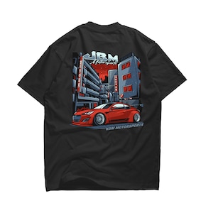 Peut inclure: T-shirt noir avec une voiture rouge conduisant dans une scène de rue de ville. La voiture porte le texte "KDM MOTORSPORTS" en bas. Le texte "JBM Motorsports" est en haut du design.