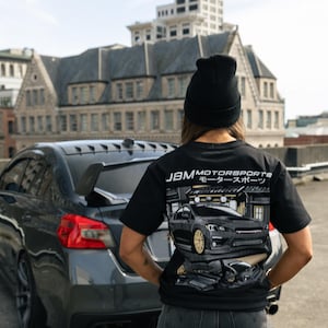 May include: Black t-shirt with a car graphic and the text "JBM Motorsports モーター スポーツ". The shirt is worn in front of a gray car. The background includes a city building.