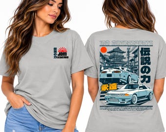 May include: A light gray short-sleeve t-shirt featuring a small JDM Legends logo on the front chest. The back displays a large graphic of two sports cars, a traditional Japanese building, a red sun, and text reading JDM MOTORSPORTS.