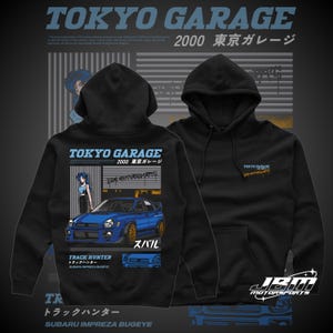 以下が含まれることがあります： 「TOKYO GARAGE 2000 東京ガレージ」の文字と青いスバルインプレッサBugEyeのイラストが描かれた黒いフード付きスウェットシャツ。「TRACK HUNTER」と「SUBARU IMPREZA BUGEYE」の文字も含まれています。