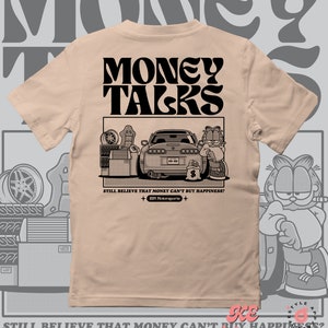 Puede incluir: Una camiseta de color beige con un estampado gráfico negro de un gato de dibujos animados sentado frente a un coche con el texto "MONEY TALKS" encima de la imagen. El texto "STILL BELIEVE THAT MONEY CAN'T BUY HAPPINESS? +JBM Motorsports+" está debajo de la imagen. El texto "www.jbmmotorsports.net Creations" está en la parte inferior de la camiseta.