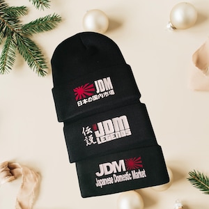 May include: Three black knit beanies stacked on top of each other. Each beanie features the letters "JDM" in white, along with Japanese text and a red sunburst design. The bottom beanie reads "Japanese Domestic Market".