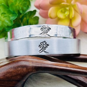 Puede incluir: Dos anillos de plata apilados, grabados con el carácter japonés que significa amor. Se exhiben sobre una escultura de mano de madera, con flores rosas y verdes en el fondo. Un símbolo de afecto.