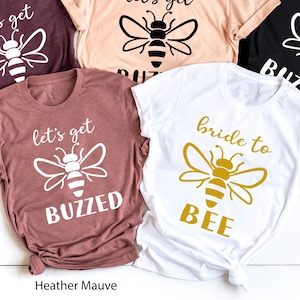 May include: Five t-shirts in various colors: maroon, peach, black, mauve, and white. Each shirt features a bee graphic with text. The text on the shirts reads "let's get buzzed" and "bride to bee".