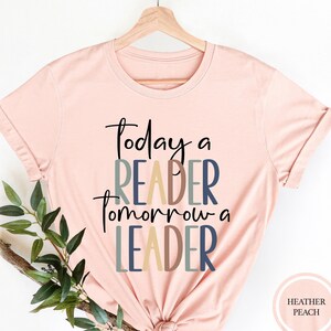 May include: A pink t-shirt with the text "Today a Reader Tomorrow a Leader" in colorful letters. The shirt is on a hanger and has a knotted bottom.
