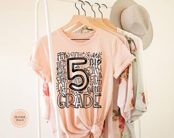 Puede incluir: Una camiseta color melocotón con un estampado gráfico negro que muestra el número 5 grande y el texto Fifth Grade rodeado de pequeños iconos escolares como un lápiz, una manzana, un globo terráqueo y una bombilla, en una percha de madera.