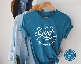 May include: A heather deep teal short-sleeve t-shirt featuring white text that reads With God all things are possible in a circular design. The shirt is displayed on a wooden hanger alongside a light blue denim shirt and a brown hat.