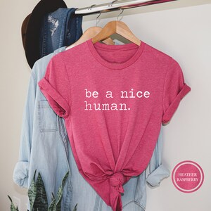 Pu&ograve; includere: Maglietta rosa con il testo "be a nice human." in lettere bianche. La maglietta &egrave; su un appendiabiti e ha un nodo sul davanti.