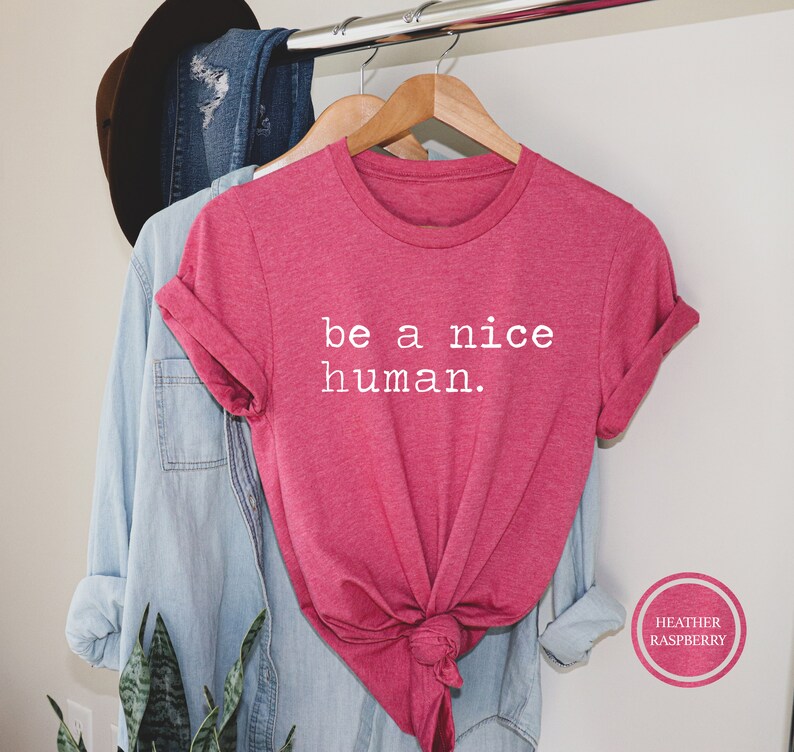 Pu&ograve; includere: Maglietta rosa con il testo "be a nice human." in lettere bianche. La maglietta &egrave; su un appendiabiti e ha un nodo sul davanti.