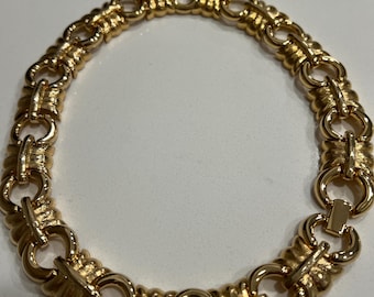 Puede incluir: Un collar dorado con un diseño de eslabones. El collar presenta una serie de eslabones interconectados, con un patrón texturizado en cada eslabón. El cierre es un cierre simple de barra y lazo. La joya se exhibe sobre una superficie blanca.
