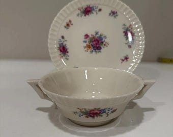 May include: Cream-colored soup bowl with two handles and a matching plate. Both pieces feature a floral pattern in pink, blue, and yellow. The plate has a scalloped edge.