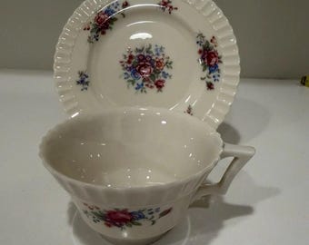 May include: Cream-colored teacup and saucer set with a floral design in red, blue, and purple. The cup has a handle and a scalloped rim, and the saucer has a matching scalloped edge. A classic tea set.