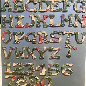 May include: A collection of wooden alphabet letters and numbers with a tropical floral pattern in green, red, and tan. The letters and numbers are arranged on a gray surface.