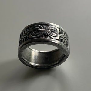 May include: A silver ring with a decorative design featuring a repeating pattern of stylized swirls and a textured band.
