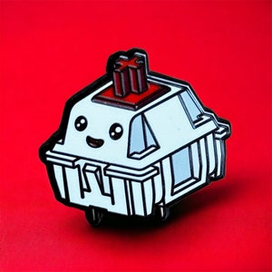 Puede incluir: Un pin de esmalte blanco con forma de tecla de teclado con una tecla roja y negra. La tecla tiene forma de cruz y la tecla tiene una cara sonriente.