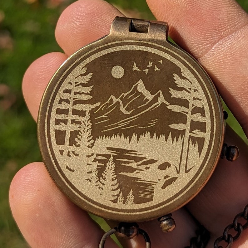 May include: A brass compass with a detailed engraved design of a mountain range and trees. The compass has a circular face with a gold finish and a black background.
