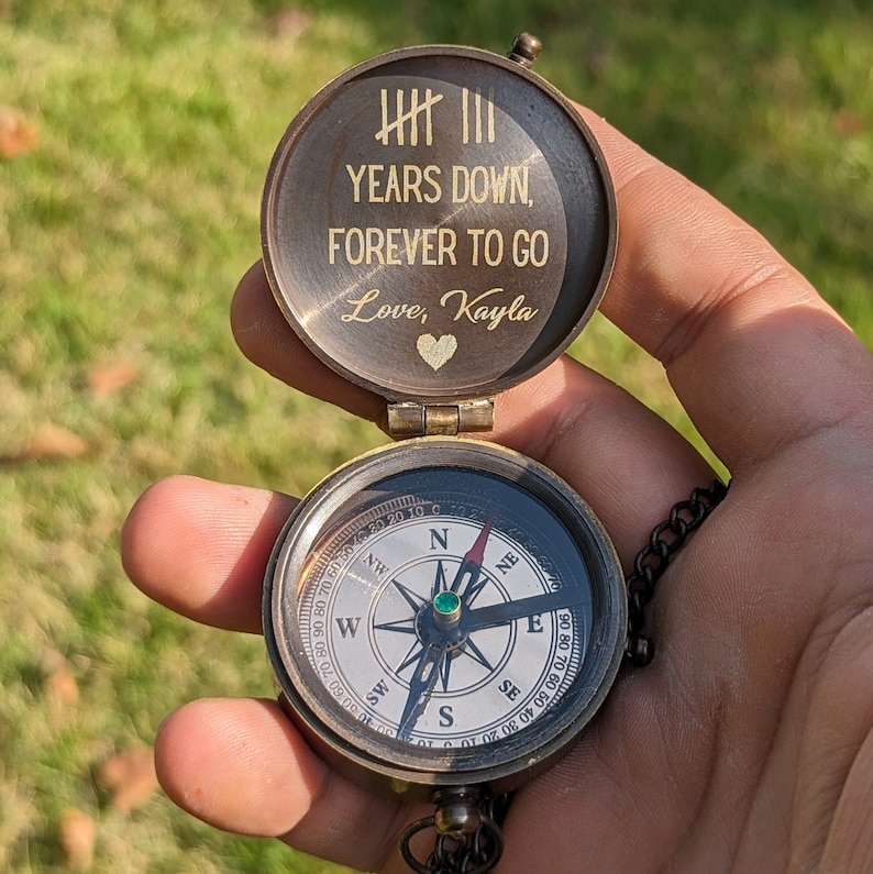 May include: A brass compass with a lid that reads "YEARS DOWN, FOREVER TO GO Love, Kayla". The compass has a green jewel in the center and a black needle.