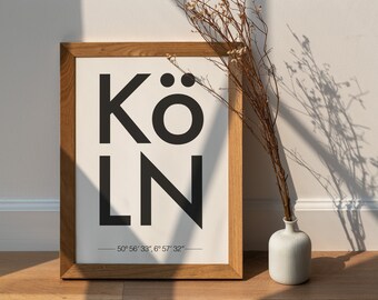 May include: A framed print with the word "KÖLN" in black text on a white background. The print also includes the coordinates "50° 56' 33", 6° 57' 32" ". The frame is made of wood and has a natural finish. A vase with dried flowers is in the foreground.