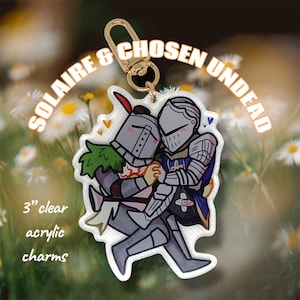 May include: Clear acrylic charm featuring two cartoon knights embracing. The charm is 3 inches in size and has a gold-colored clasp. Text on the charm reads "SOLAIRE & CHOSEN UNDEAD". The knights are in grey armor with accents of red, blue, and green.