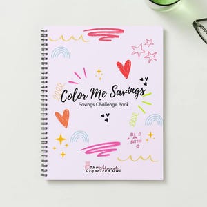 May include: A spiral-bound notebook with a light pink cover featuring the text "Color Me Savings" and colourful doodles. The notebook includes hearts, stars, and rainbows. The words "Savings Challenge Book" are also visible.
