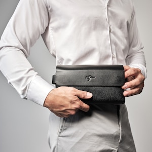 Puede incluir: Un clutch de cuero negro con un logotipo de canguro. El clutch lo lleva una persona que lleva una camisa blanca abotonada y pantalones grises claros.