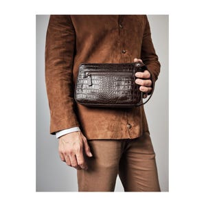 May include: A dark brown, crocodile-textured clutch with a zipper closure. The rectangular bag has a wrist strap and is held against a person wearing a brown suede jacket and tan pants.