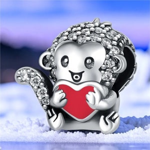 Puede incluir: Un charm de plata con forma de mono que sostiene un corazón rojo. El mono está decorado con pequeños cristales blancos.