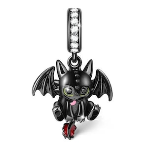 Puede incluir: Un charm de dragón negro con cola roja y ojos verdes. El charm tiene una parte superior plateada con cristales transparentes. El dragón tiene alas detalladas y está sentado.