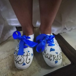 May include: A pair of white sneakers with blue ribbons tied around the ankles. The left shoe says "Just" and the right shoe says "Married".