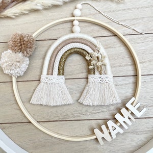 Könnte beinhalten: Ein beige-goldener Makramee-Regenbogen-Wandbehang mit einem Holzreifen und dem Wort "NAME" in weißen Buchstaben. Der Regenbogen hat einen weißen Spitzenbesatz und beige Quasten.